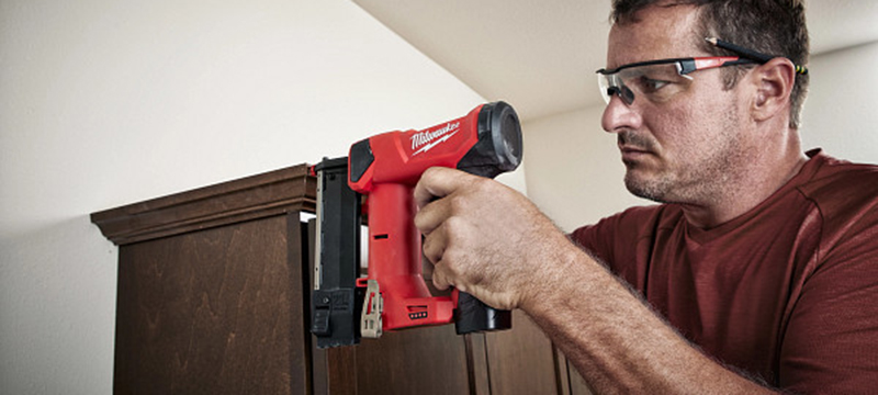 A worker uses a Milwaukee M12 23 Gauge Cordless Pin Nailer to attach trip to the top of a cabinet.