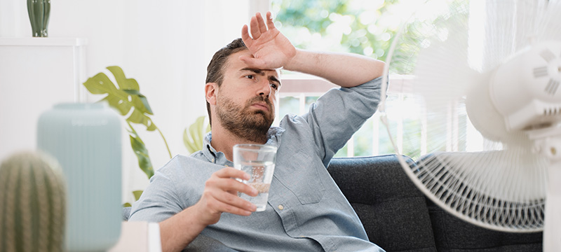 A man tries to deal with the heat with a cold glass of water and a fan.