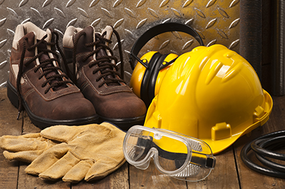 Personal protective equipment, such as work boots, hard hat, ear muffs, goggles, and leather work gloves sit on a workbench.