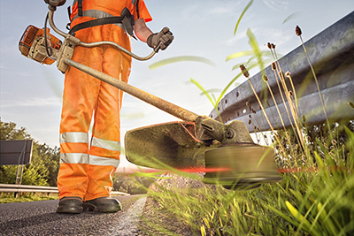 A gas-powered string trimmer is used to trim grass along a highway.