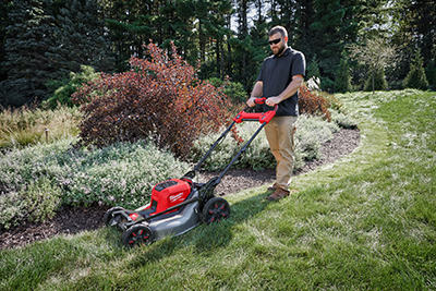 A man pushes the Milwaukee M18 FUEL Self-Propelled Dual Battery Mower down a decline around a flower bed.