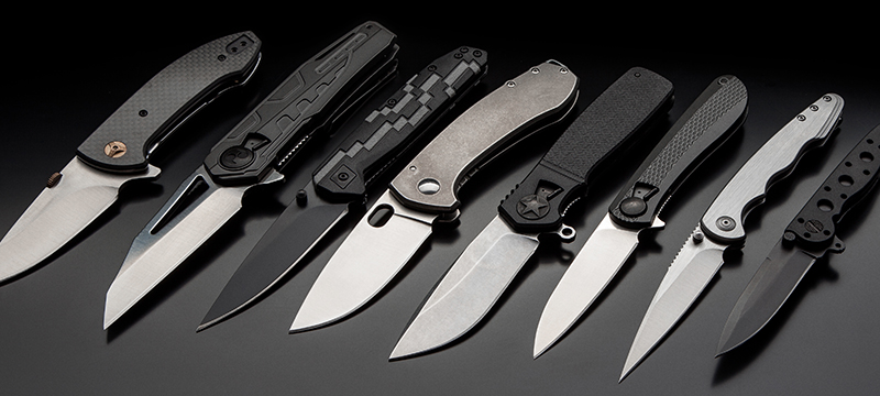 Several everyday carry pocket knives lined up on a black table.