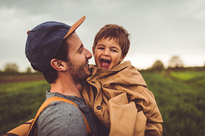 A dad wearing a denim and canvas hat holds his son.