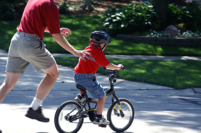 A dad in khaki shorts and calf socks helps his son ride a bike.