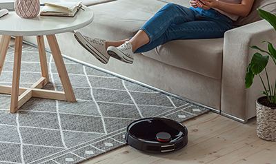 A woman lifts her legs so a robotic vacuum can clean under her feet.