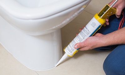 Silicone is applied to the bottom of a toilet with a caulk gun.