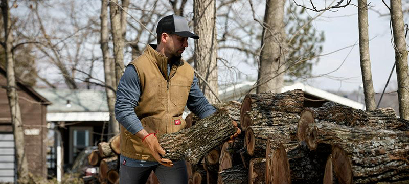 A man wearing a brown Milwaukee Heavy Duty Sherpa-Lined Vest stacks wood.