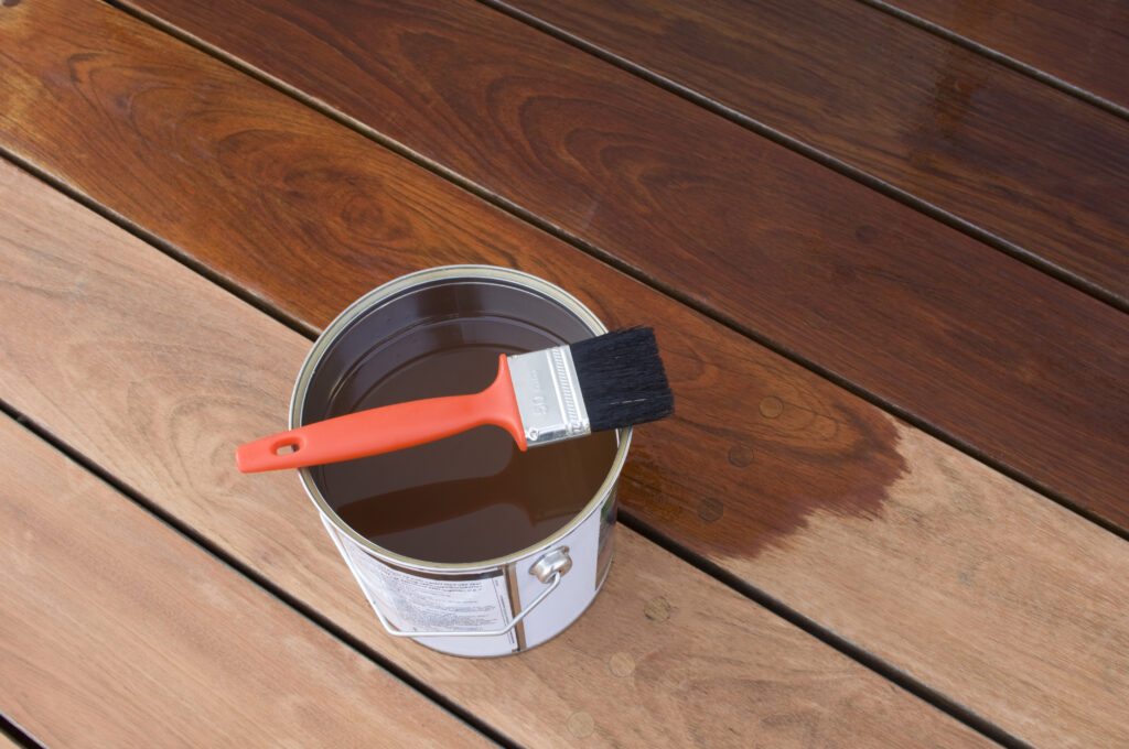 Container of brown wood stain with paint brush set on top of a wooden planks. 