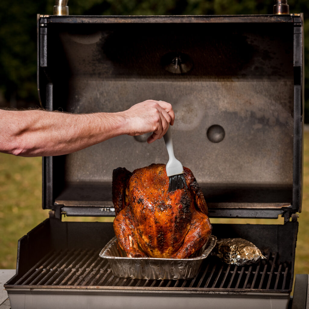 Person basting turkey that is cooking on a grill. 