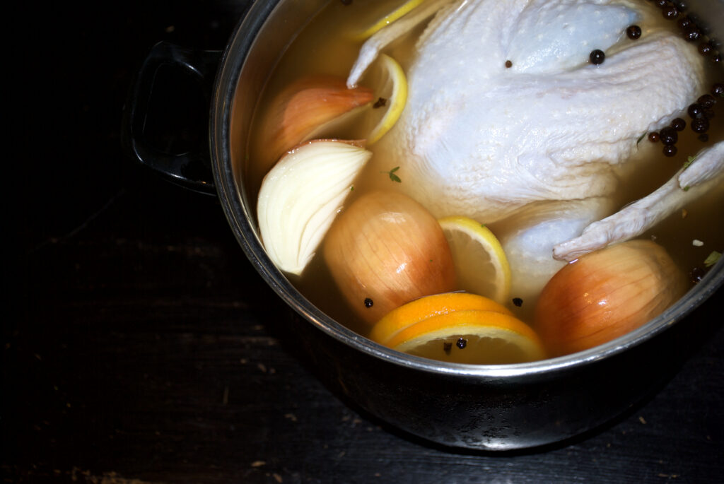 Raw turkey brining in metal pot with liquid, onions, lemons, and peppercorns. 